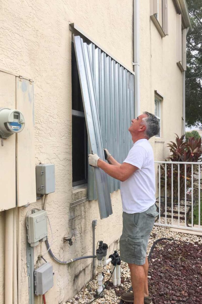 A man in a white t-shirt and cargo shorts installs corrugated metal storm shutters over a window on the side of a stucco house - www.frankslaughterinsurance.com